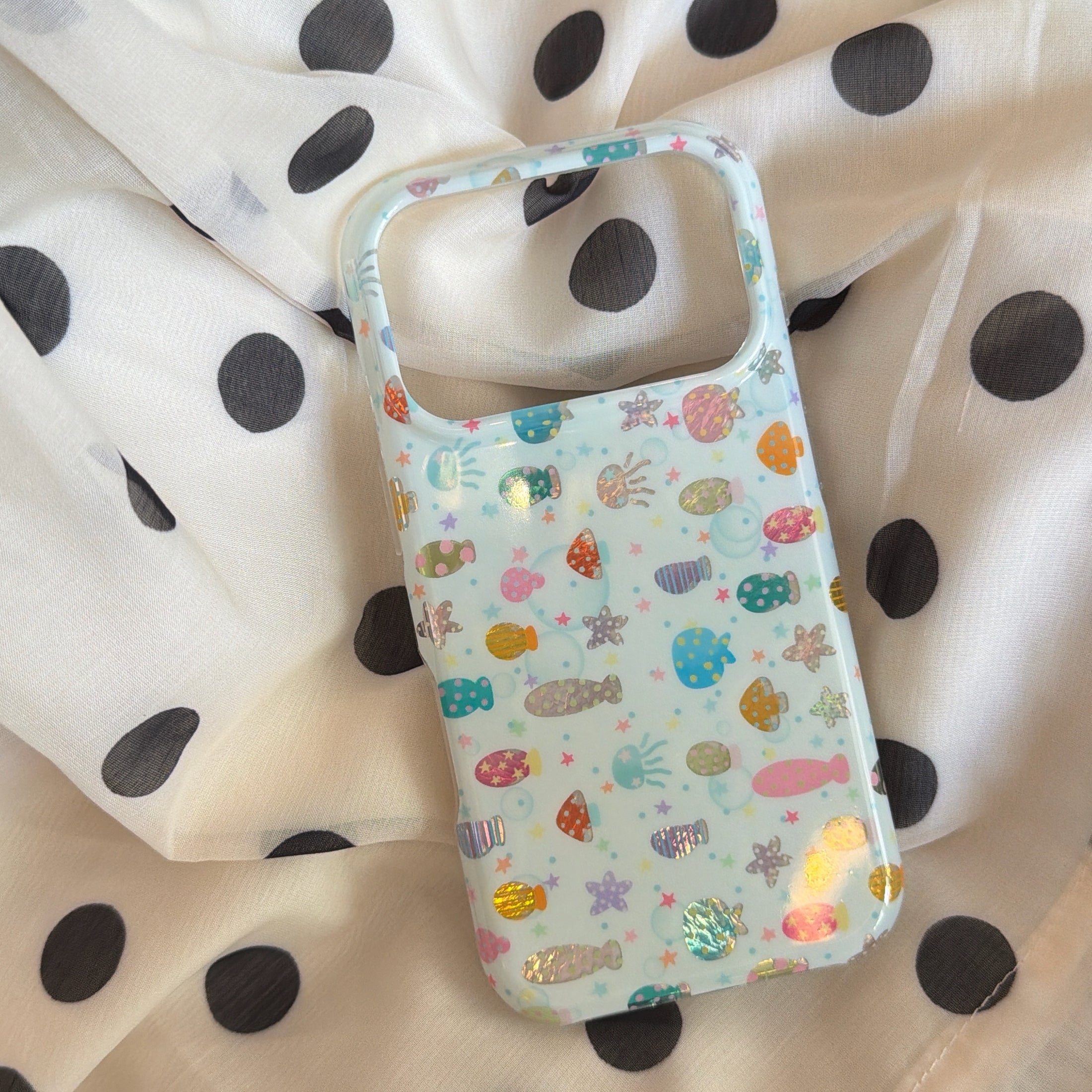 An iridescent holographic ocean fish phone case showing glossy reflections on a white polka dot fabric.