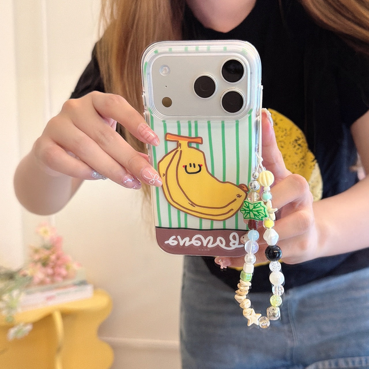 A girl holding a clear iPhone case with a cute smiling cartoon banana on a green striped background, featuring a colorful beaded phone charm.