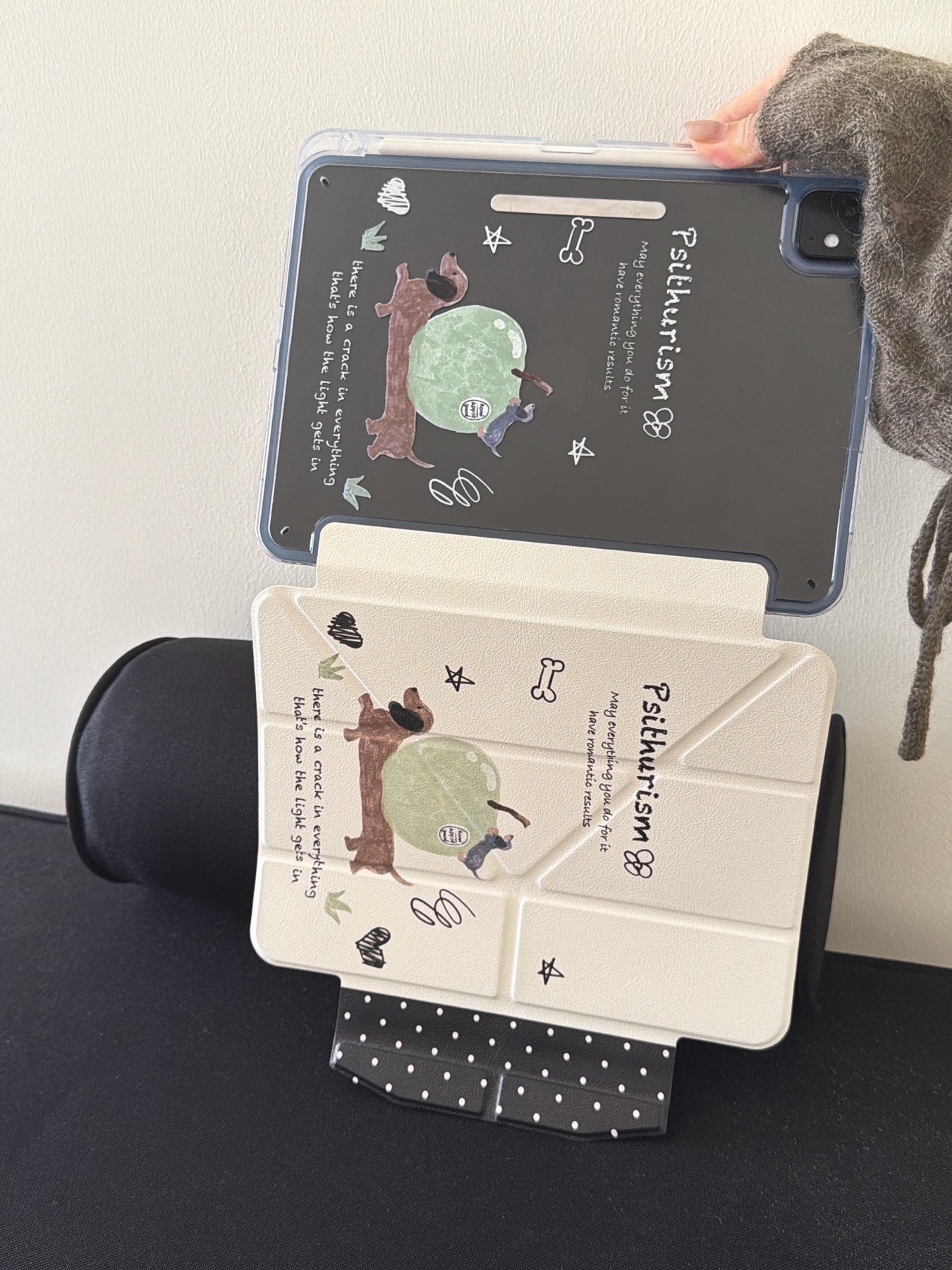 An open view of a kawaii doodle ipad smart cover leaning against a black cylindrical cushion, displaying both the printed transparent back shell and the fully extended cream origami front cover.