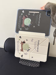 An open view of a kawaii doodle ipad smart cover leaning against a black cylindrical cushion, displaying both the printed transparent back shell and the fully extended cream origami front cover.