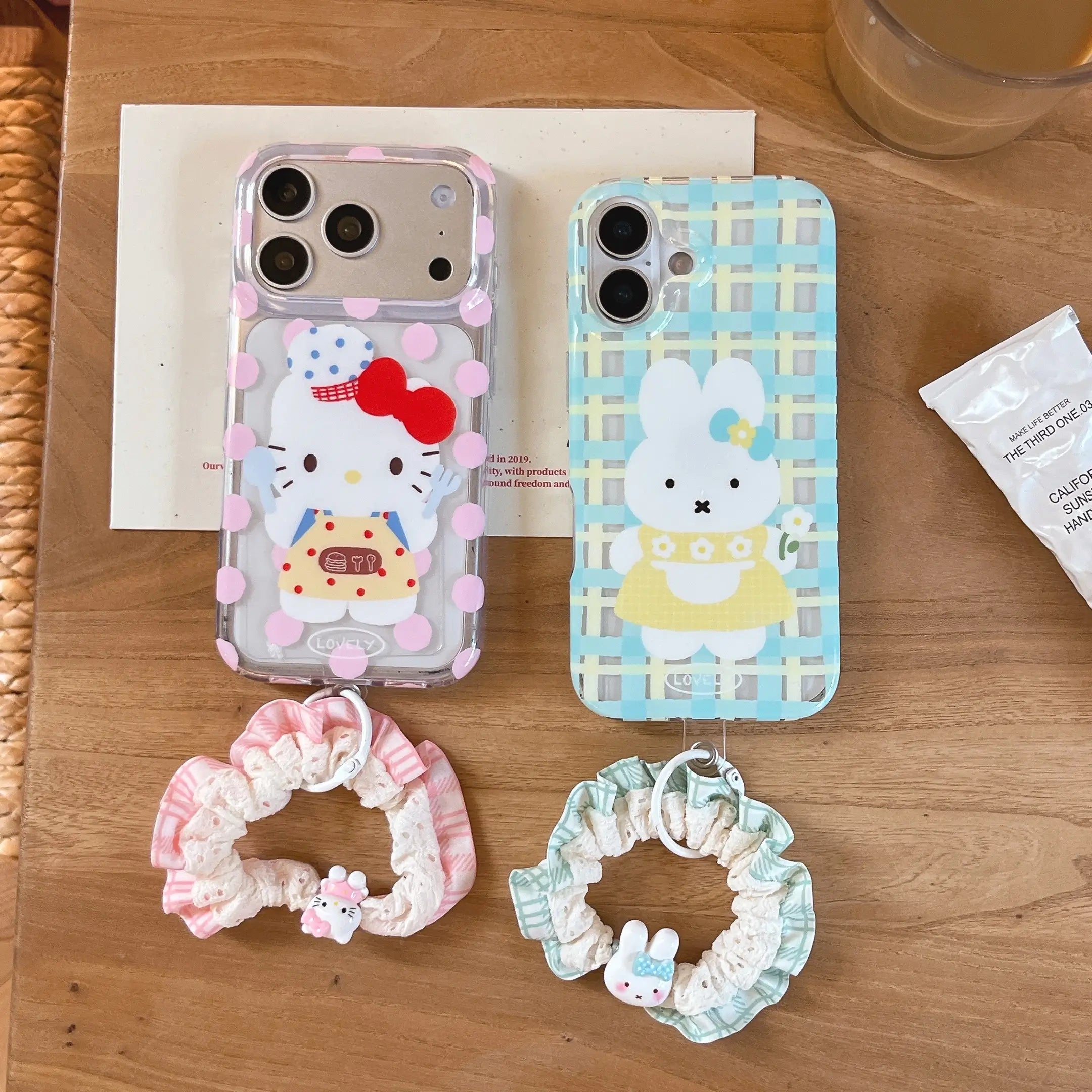 Two kawaii iPhone cases on a wooden table: one pink polka dot kitty case and one blue plaid bunny case with matching scrunchie wrist straps.