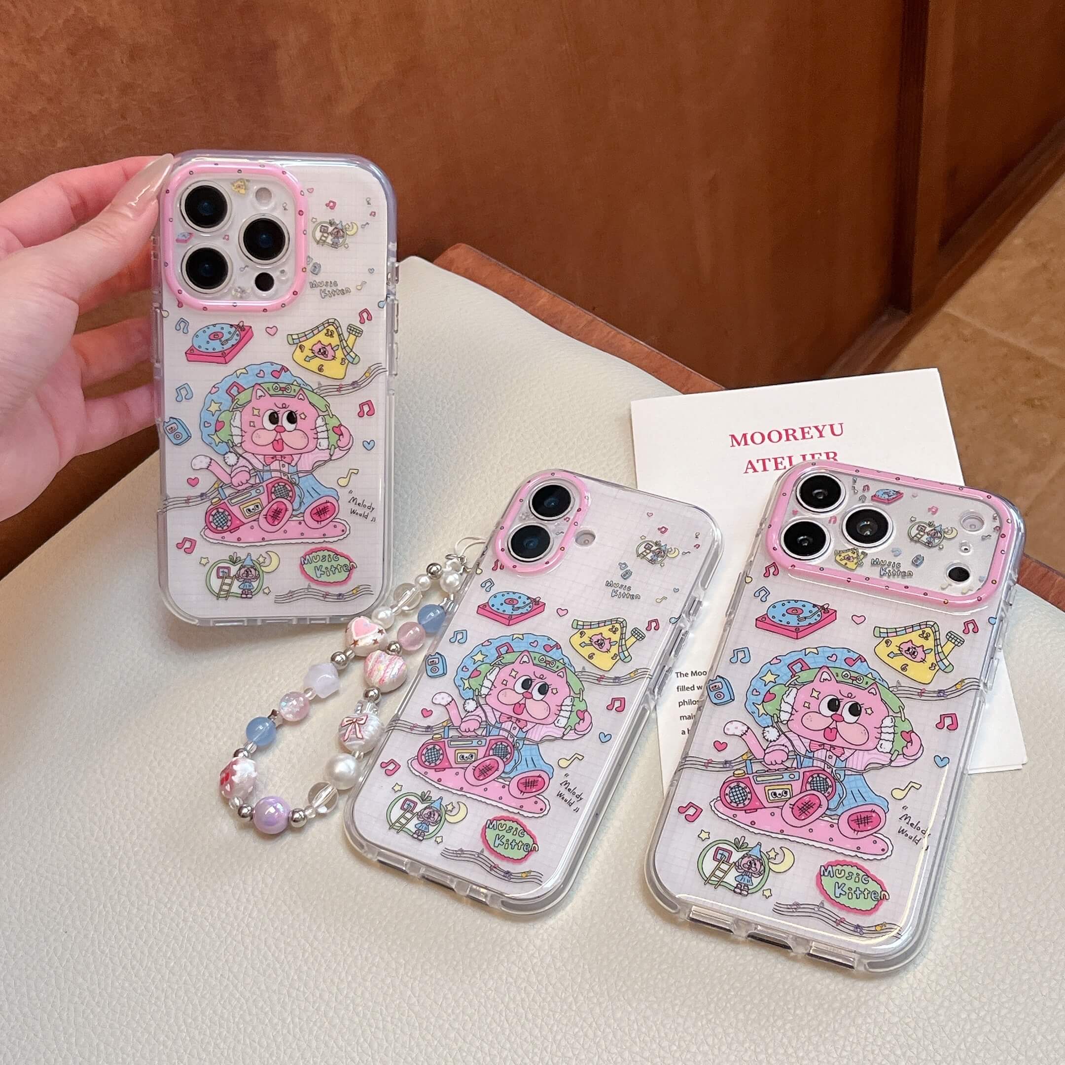 Three kawaii pink music kitten phone cases displayed together, one with a beaded wrist strap.