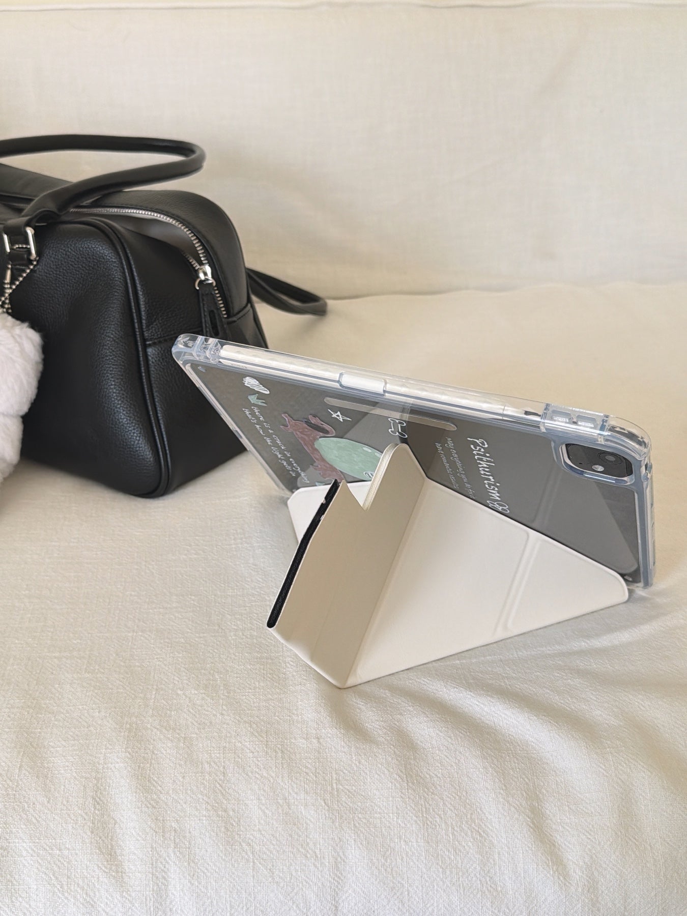 A kawaii origami tablet cover propped up horizontally on a cream-white fabric sofa next to a black leather handbag, utilizing its geometric folding stand.
