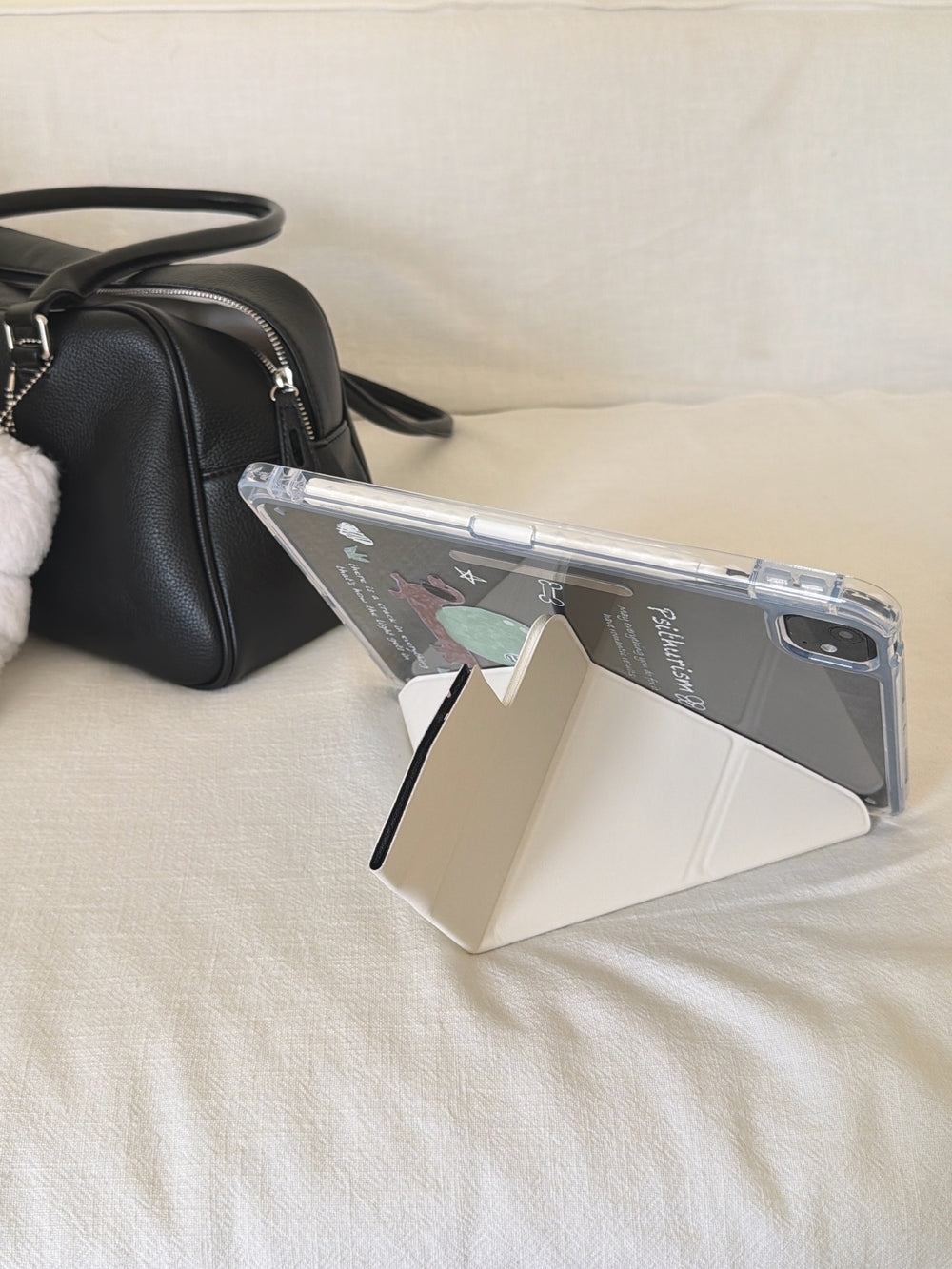 A kawaii origami tablet cover propped up horizontally on a cream-white fabric sofa next to a black leather handbag, utilizing its geometric folding stand.