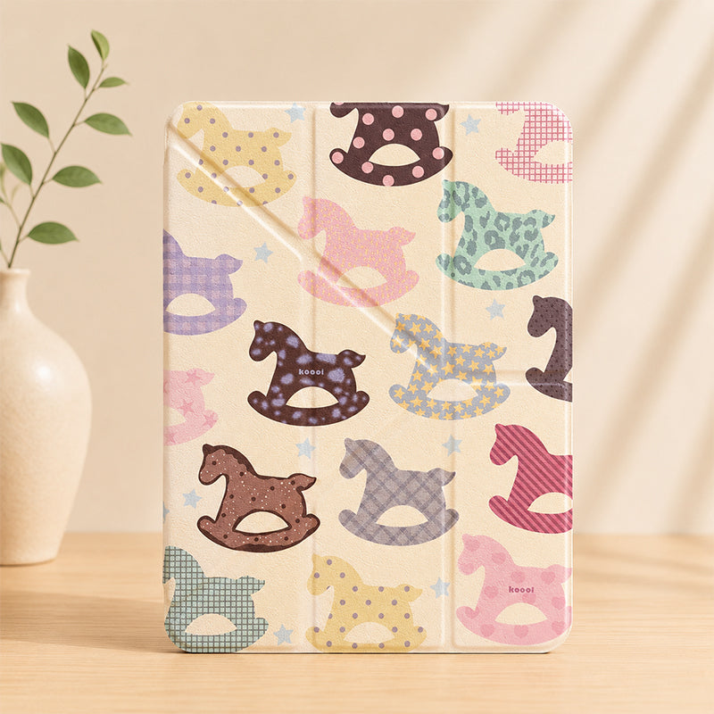 An upright lifestyle studio shot of a closed cream-colored kawaii rocking horse ipad case standing on a wooden table, next to a minimal beige ceramic vase with green leaves under warm shadow.