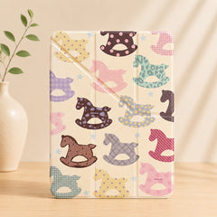 An upright lifestyle studio shot of a closed cream-colored kawaii rocking horse ipad case standing on a wooden table, next to a minimal beige ceramic vase with green leaves under warm shadow.