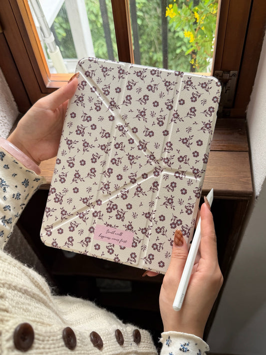 A handheld close-up of a kawaii split magnetic ipad case featuring an all-over small floral print on a cream background held near a wooden window.