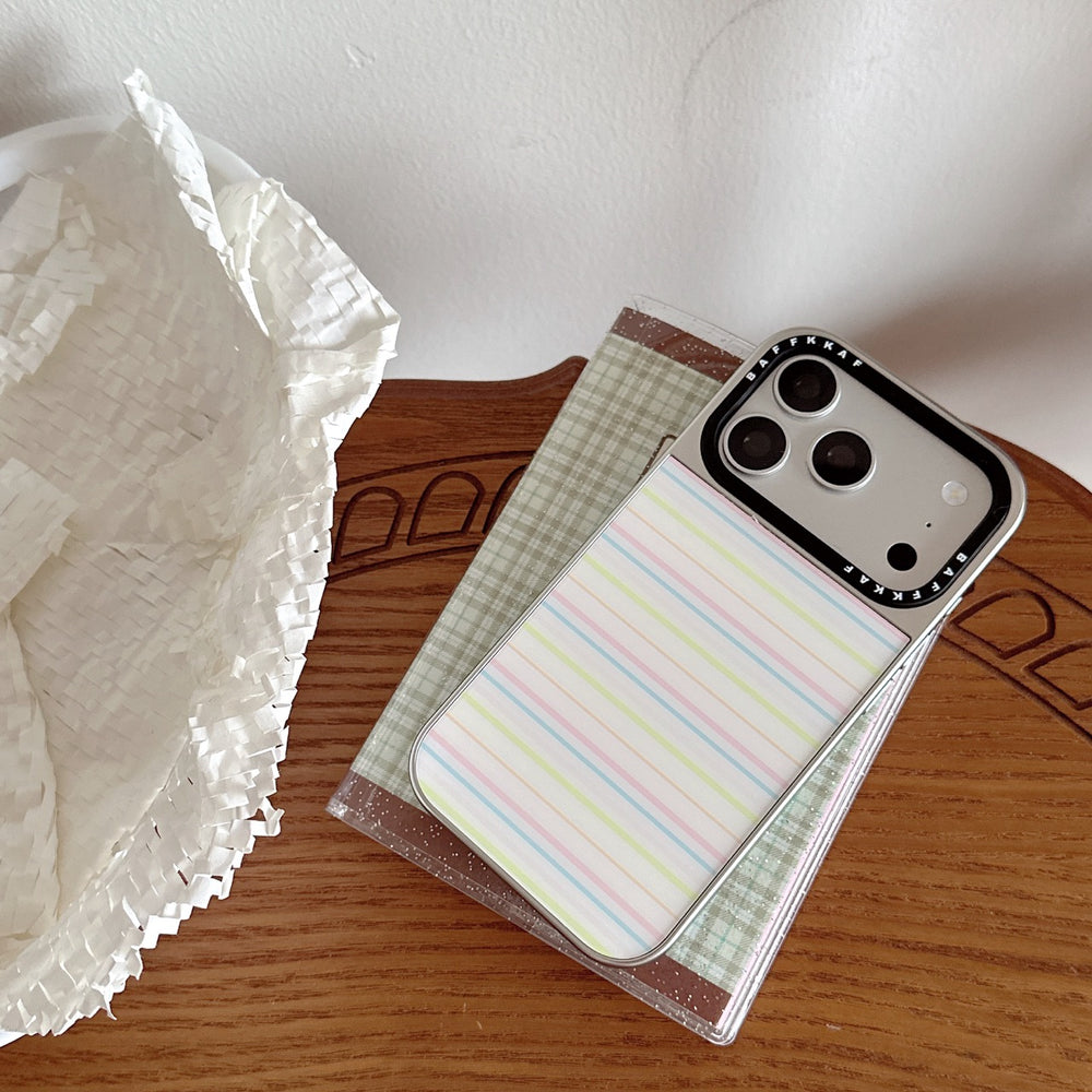 A silver Korean style striped iPhone case lying on a plaid notebook on a wooden table.
