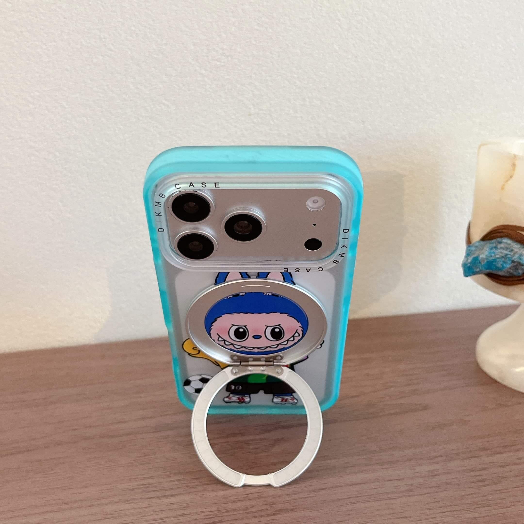 A cute Labubu soccer iPhone case standing vertically on a wooden table using its silver metal ring kickstand.