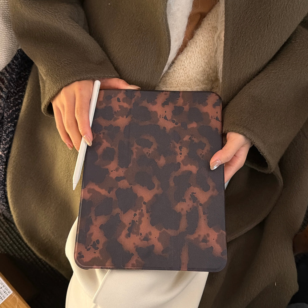 Close-up of a person holding a closed leopard print tri fold tablet case on their lap while gripping a white Apple Pencil, highlighting the sleek dark academia tech aesthetic.