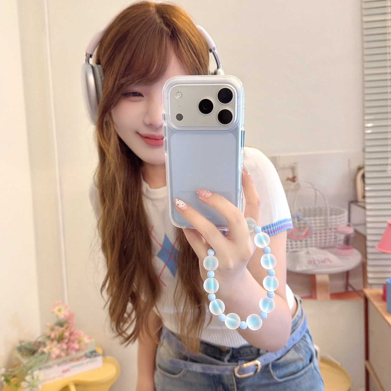 Girl with headphones posing for a mirror selfie showcasing a light blue pastel phone case with a frosted beaded lanyard.