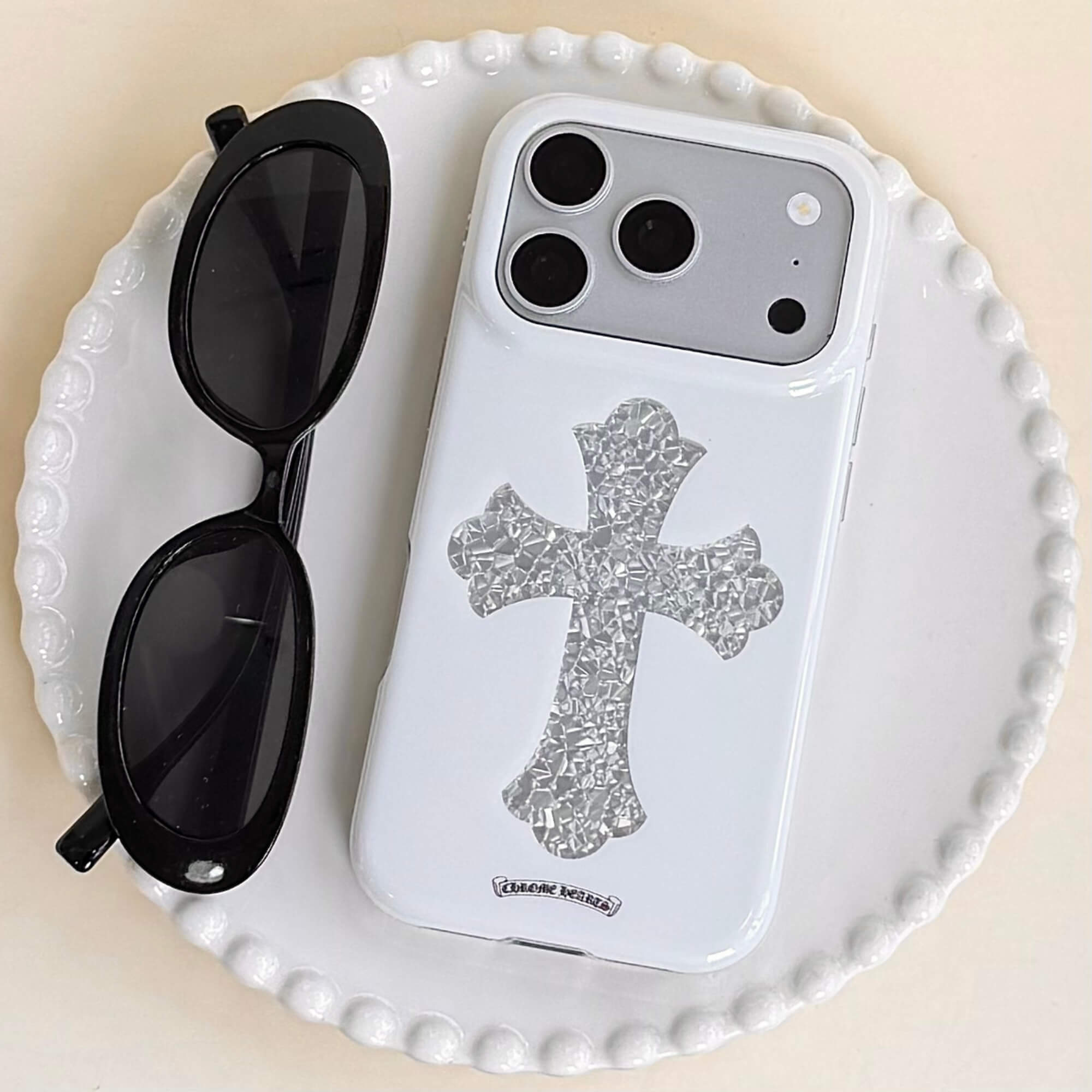 A luxury gothic silver cross phone case resting on a white ceramic plate beside black sunglasses.