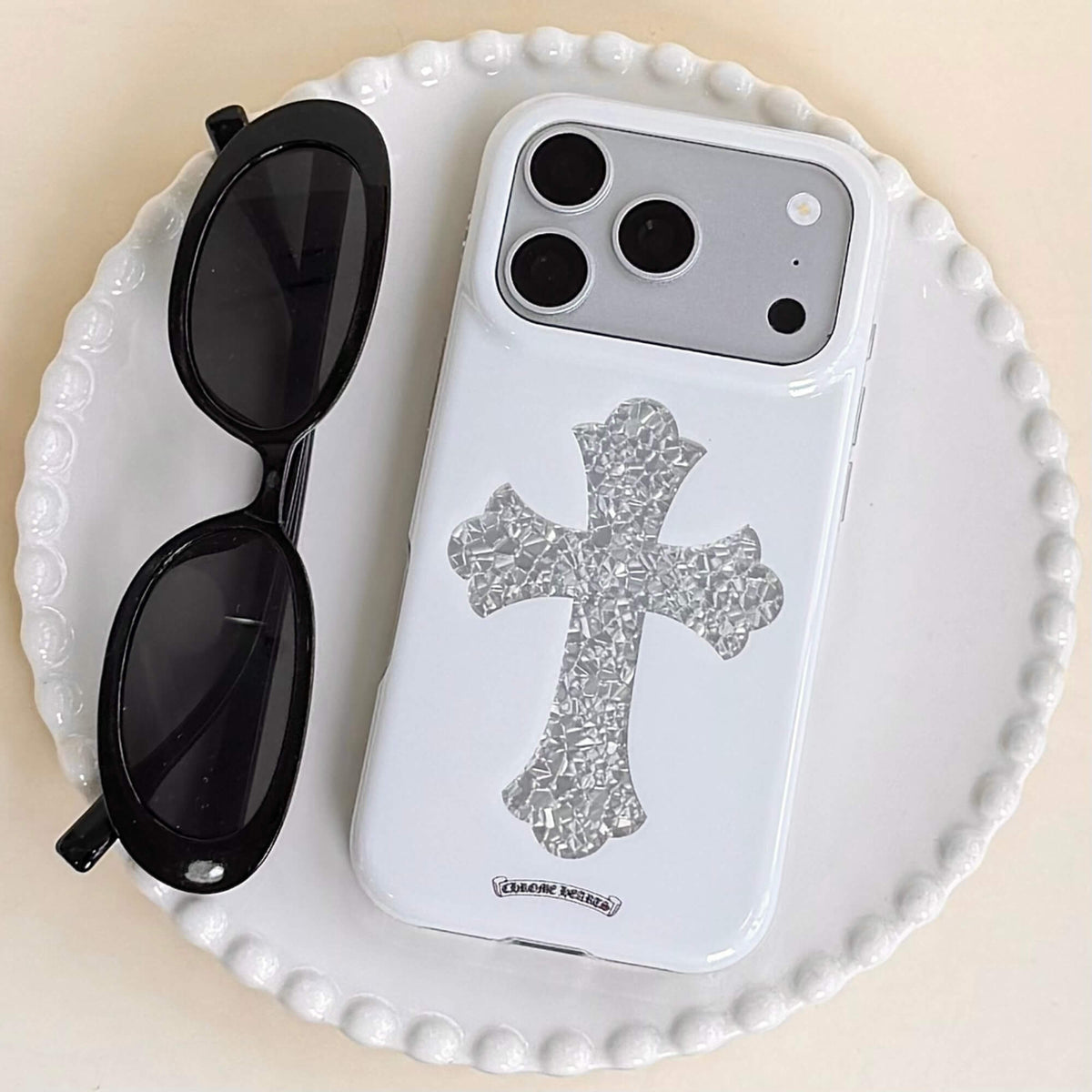 A luxury gothic silver cross phone case resting on a white ceramic plate beside black sunglasses.