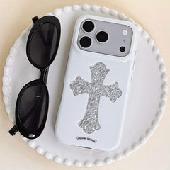 A luxury gothic silver cross phone case resting on a white ceramic plate beside black sunglasses.