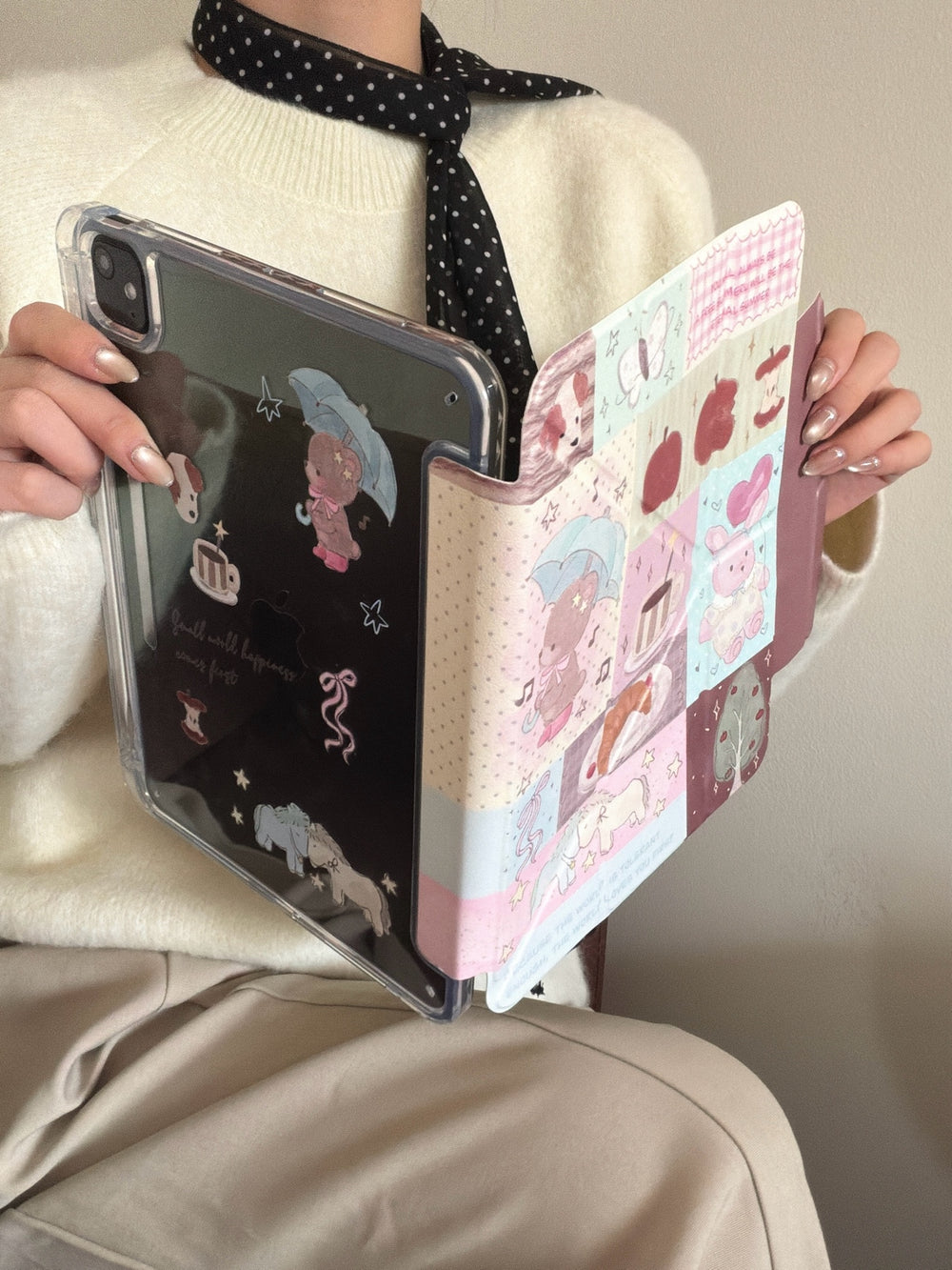 A model in a cream sweater holding an open aesthetic iPad case, displaying the magnetic detachable clear back with kawaii prints.