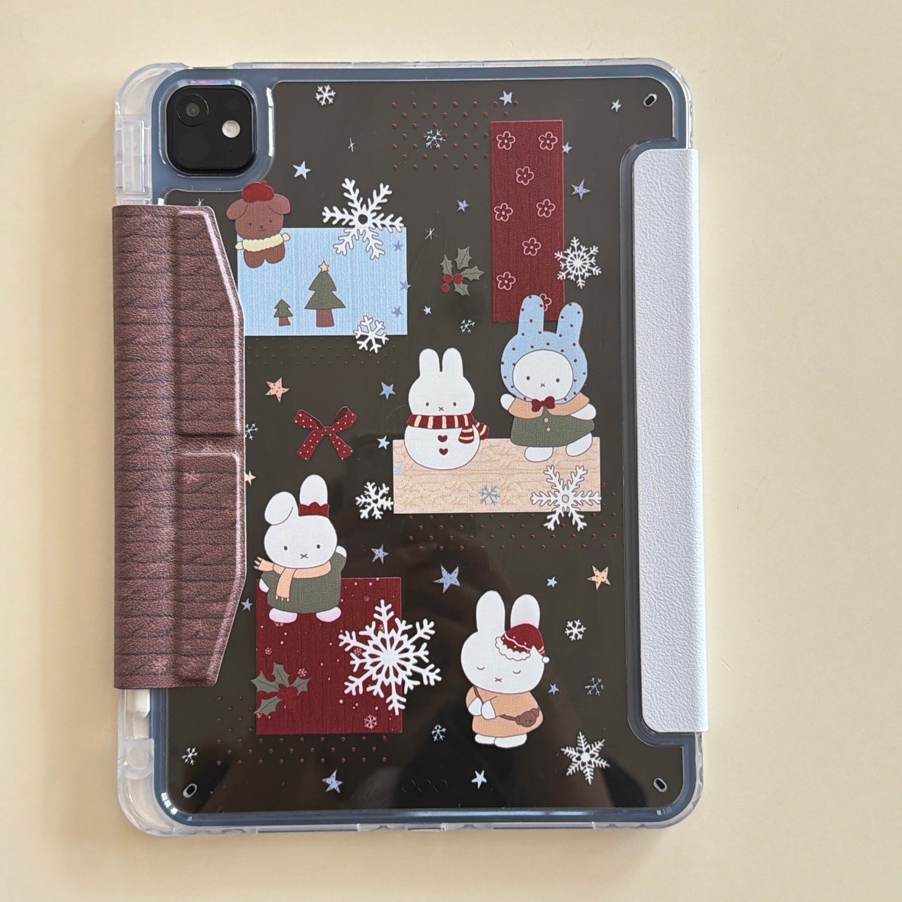 An aesthetic clear back Miffy iPad case flat on a desktop, showcasing the full layout of the cute winter bunny prints, protective pencil holder slot, and clear bumper edges.