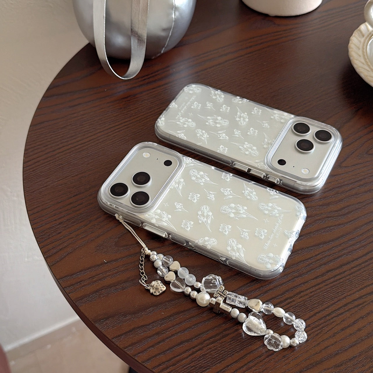 Two transparent phone covers with white flower sketches and a crystal beaded lanyard on a dark wooden table.