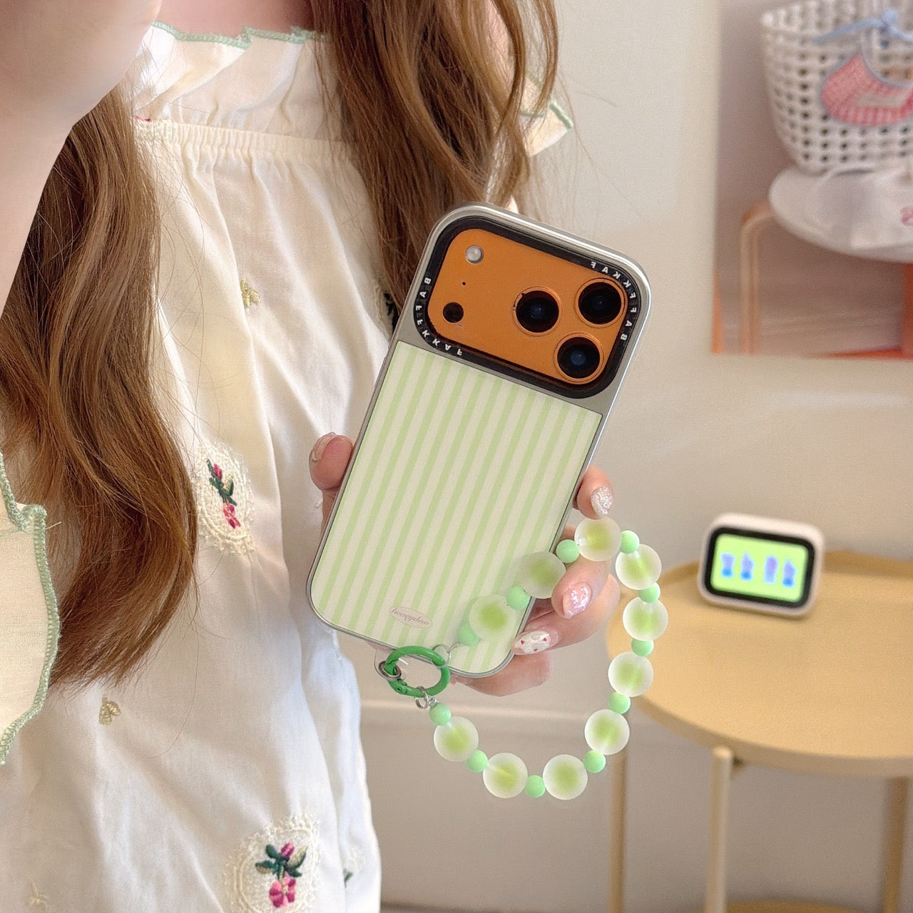 A girl holding a mint green striped iPhone case with a vibrant orange camera base and a green beaded wrist strap.