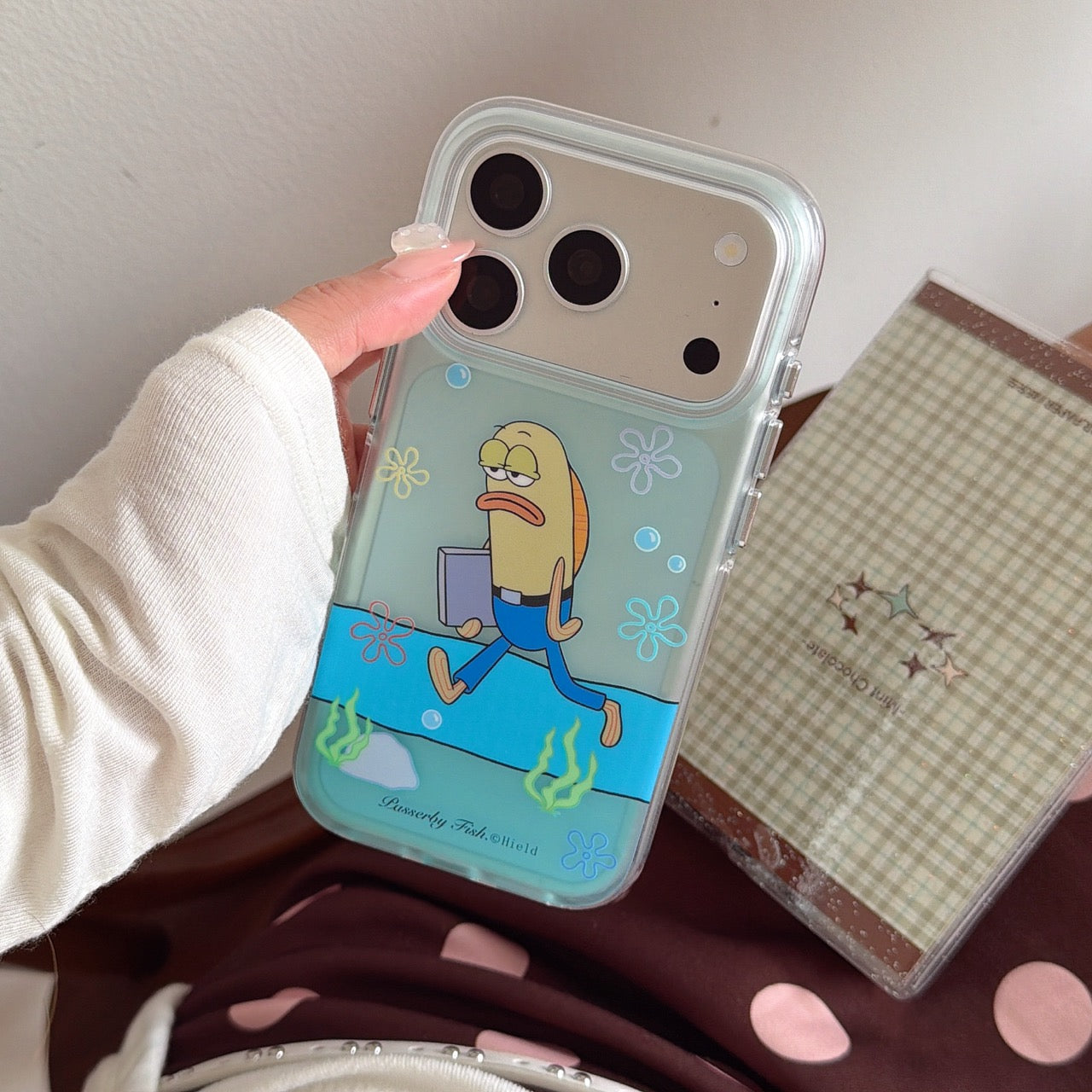 A close-up of a mint green clear phone case featuring a walking cartoon fish character held over a checkered notebook.