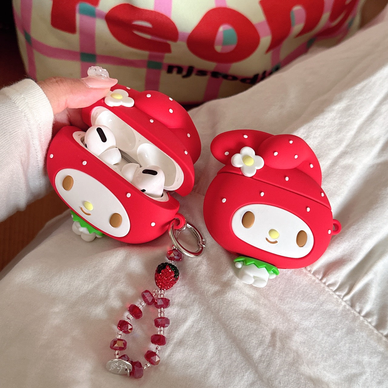 An open red My Melody strawberry AirPods case revealing white wireless earbuds inside next to a closed case.
