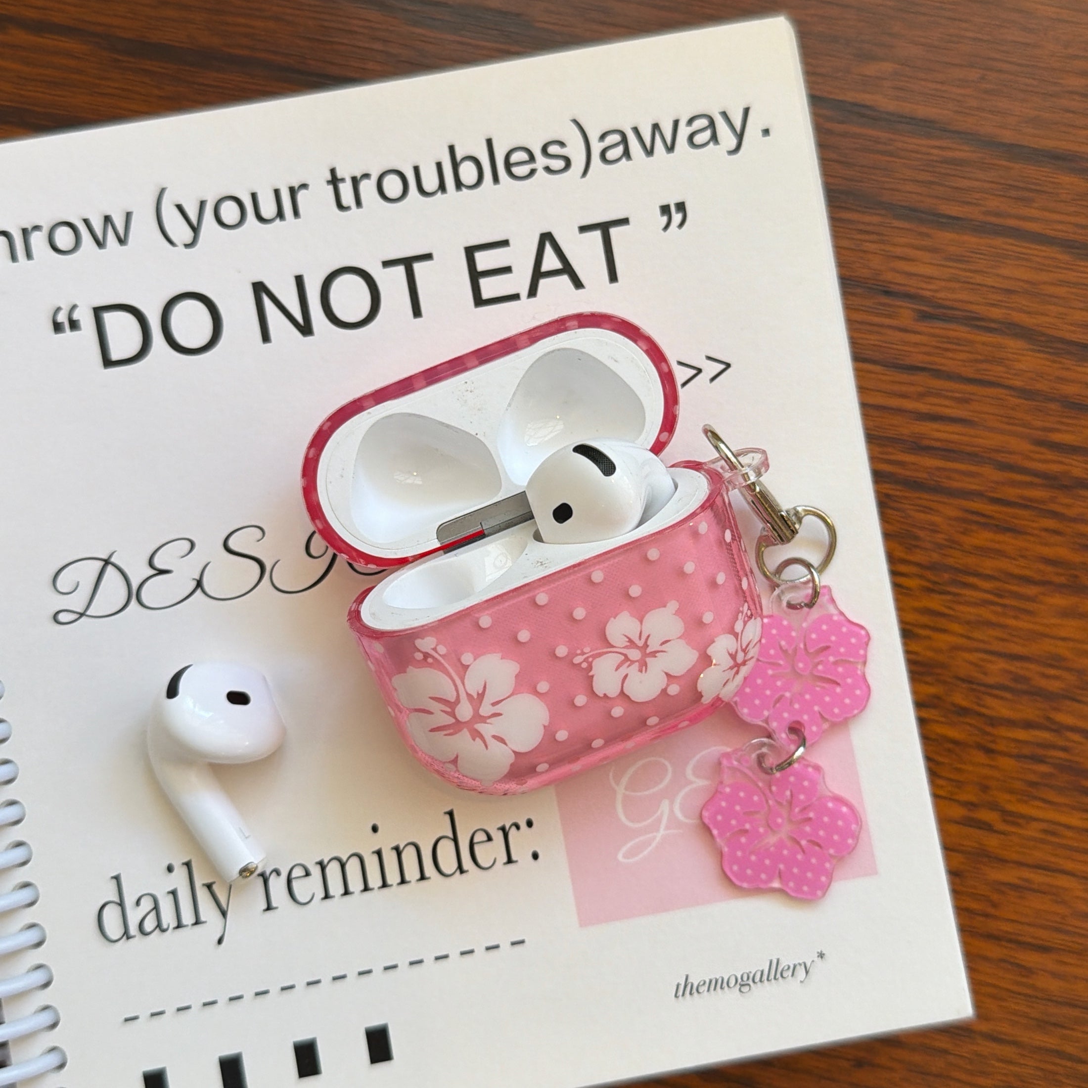 An open clear pink flower AirPods case with a white earbud resting on a white aesthetic daily reminder notebook.
