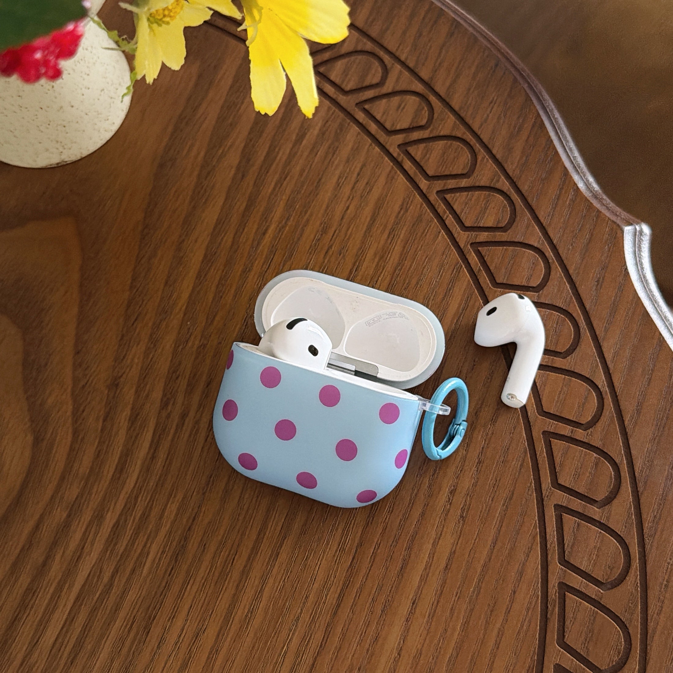An open light blue polka dot AirPods Pro case with one earbud inside and one resting next to it on a carved wooden table.