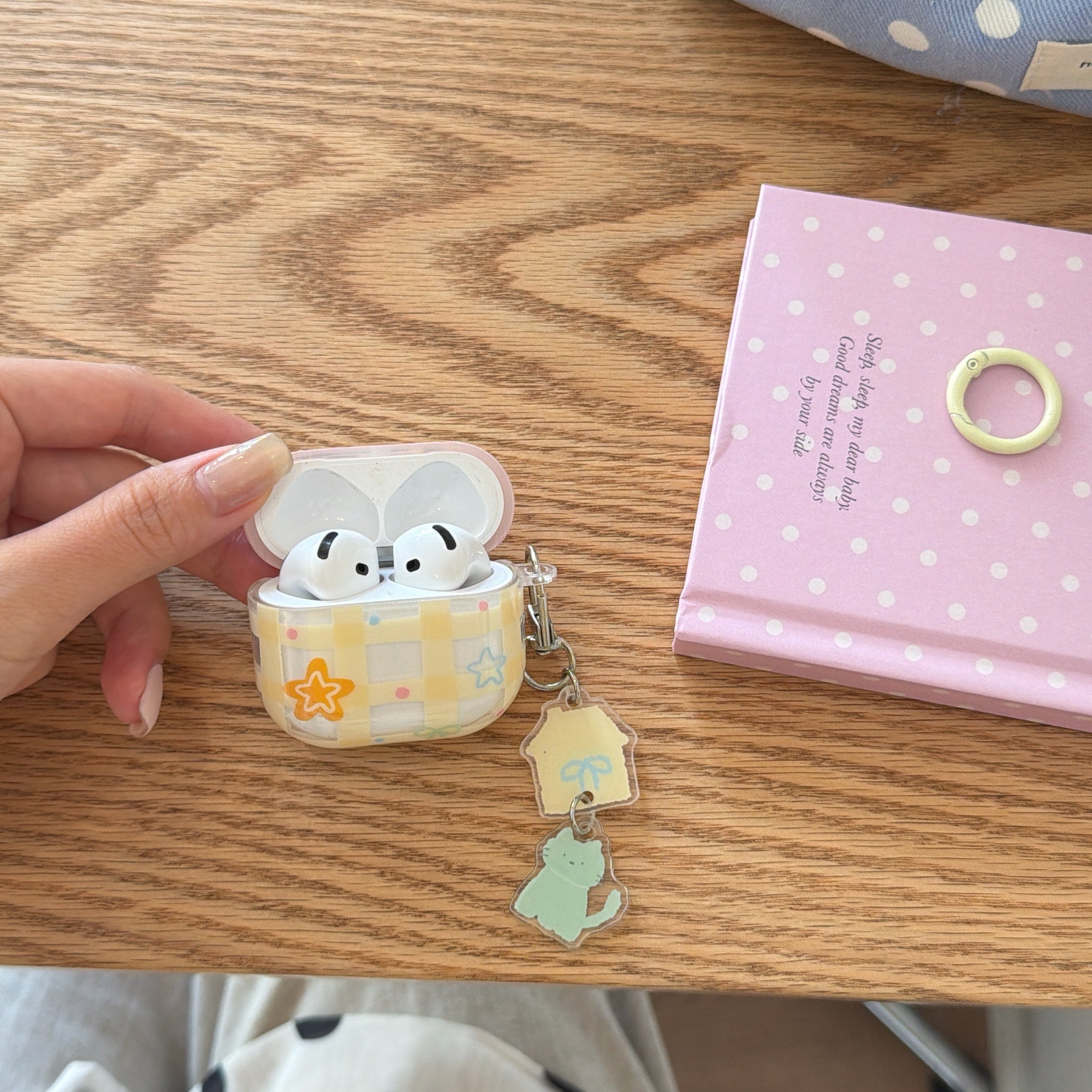 An open yellow plaid AirPods case with white earbuds inside lying on a wooden desk next to a pink notebook.