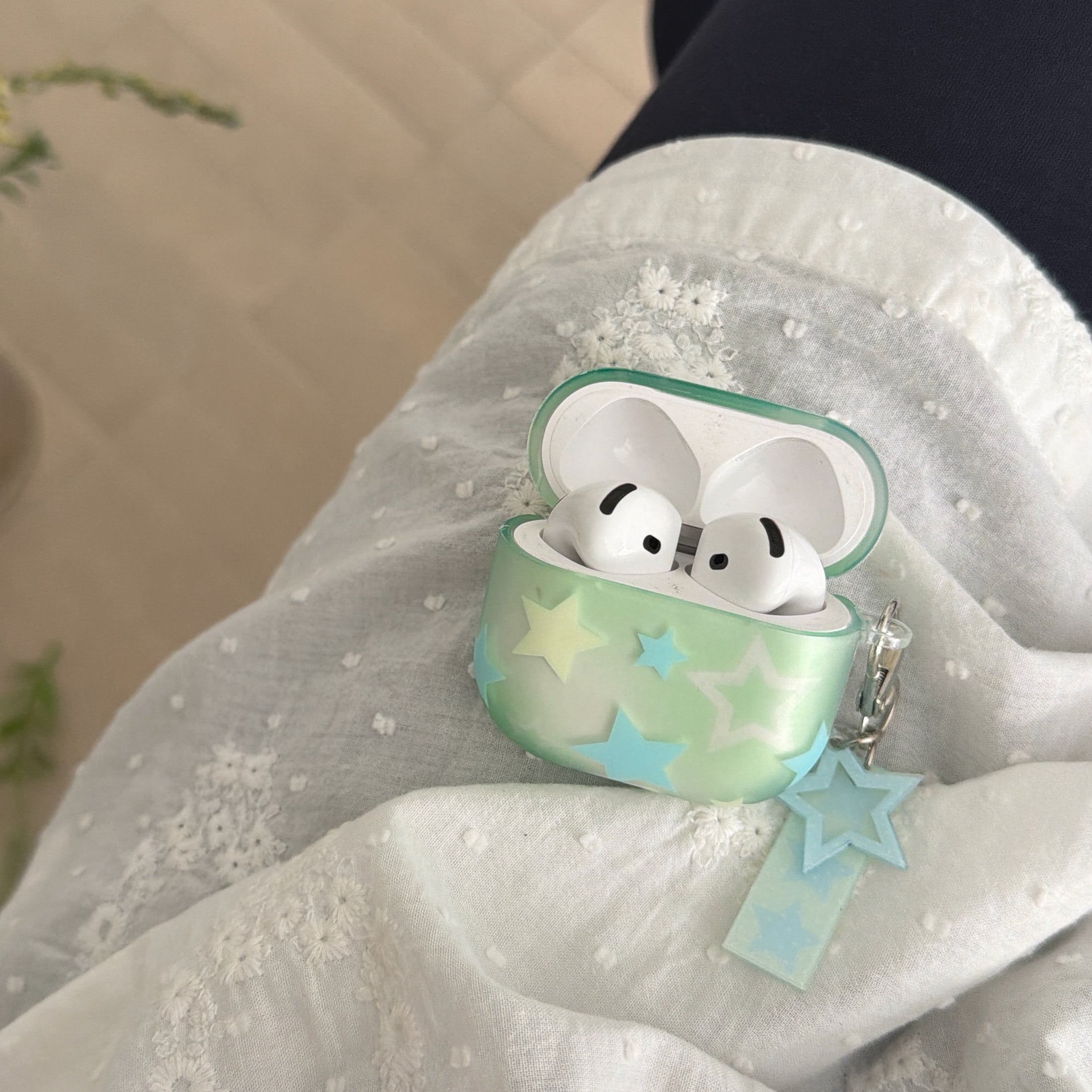 An open starry mint green AirPods case with white earbuds inside, resting on a white embroidered fabric.