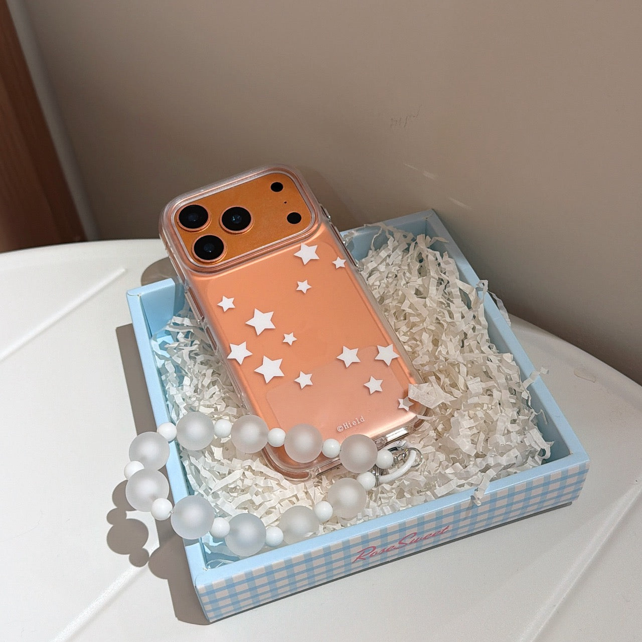An orange star-themed aesthetic iPhone case with a beaded strap packaged inside a blue checkered gift box.