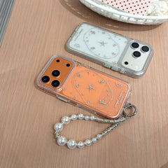 Two glittery iPhone cases in orange and metallic silver displayed flat on a wooden desk, one equipped with a luxury pearl charm strap.