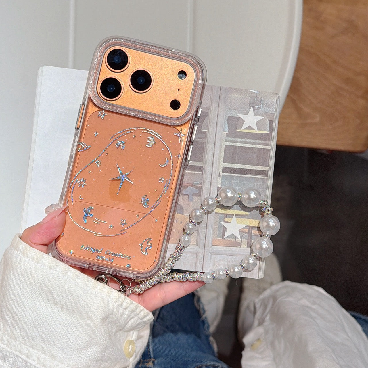 A hand holding an orange glitter galaxy phone case with a contrast metallic camera deck and a delicate pearl bead wristlet lanyard.