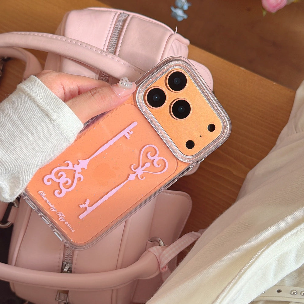 A hand holding an orange metallic charming key phone cover against a pastel pink quilted leather handbag.