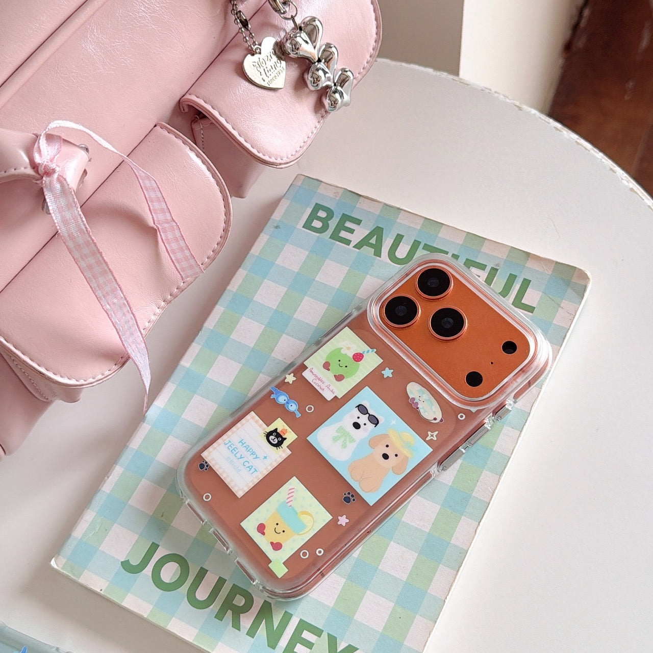 An orange holiday sticker themed phone case resting on a plaid book next to a pastel pink handbag.