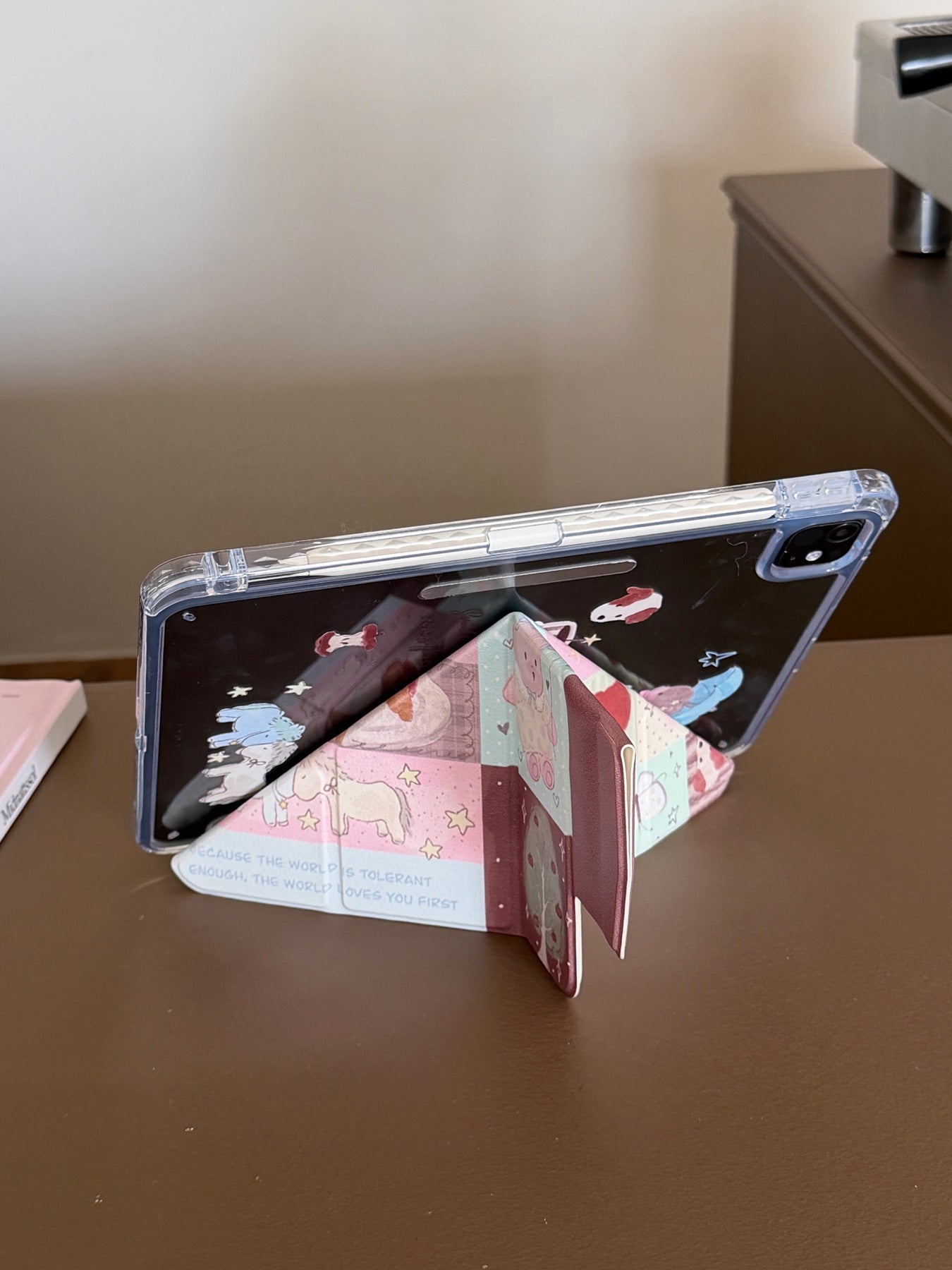 An iPad supported horizontally in landscape mode by the Y-shaped origami folding stand of a kawaii patchwork case on a brown desk.