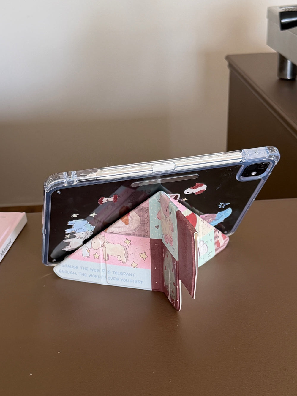 An iPad supported horizontally in landscape mode by the Y-shaped origami folding stand of a kawaii patchwork case on a brown desk.