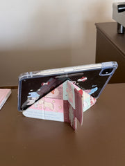 An iPad supported horizontally in landscape mode by the Y-shaped origami folding stand of a kawaii patchwork case on a brown desk.