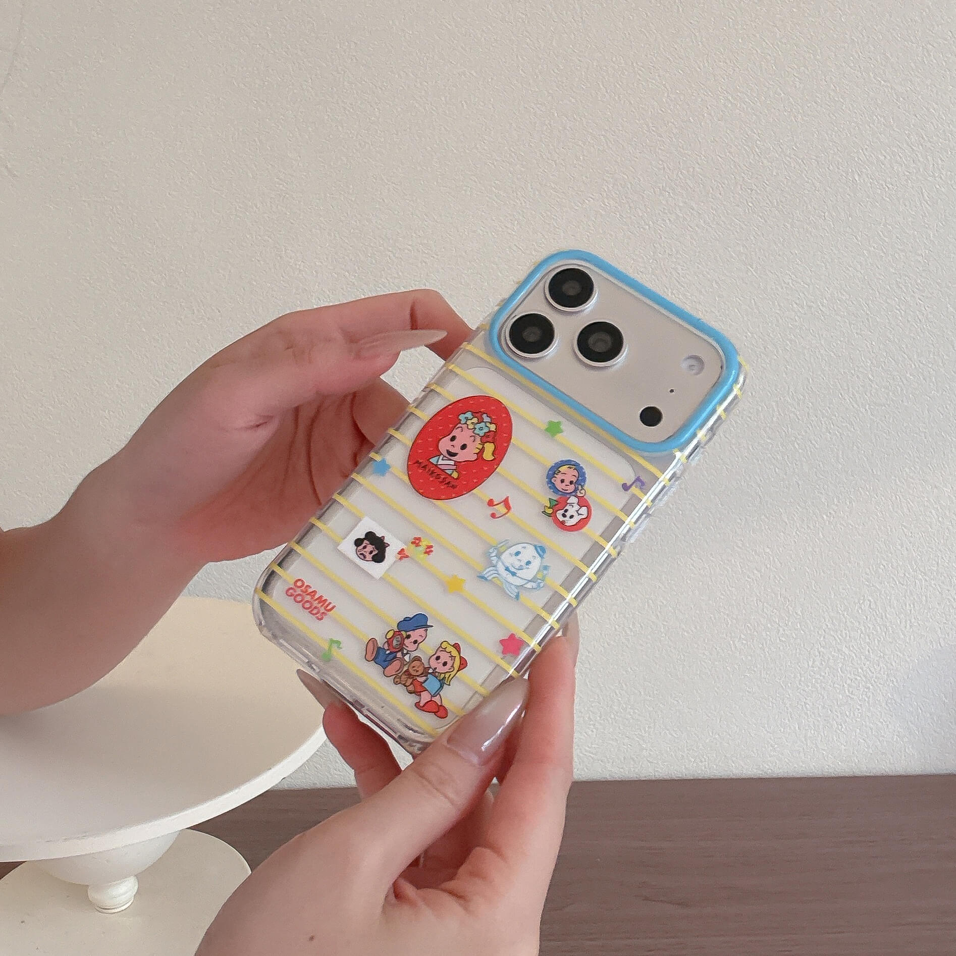 A close-up of a person holding a clear Osamu Goods iPhone case with yellow stripes and a blue camera ring.