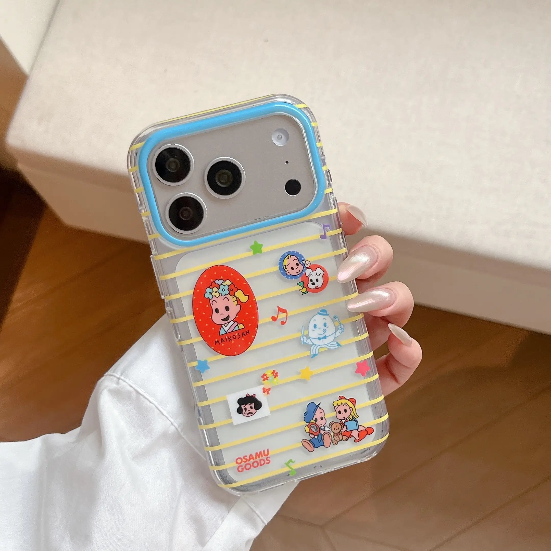 A hand holding a clear Osamu Harada striped iPhone case showing cute cartoon graphics and a blue lens protection border.