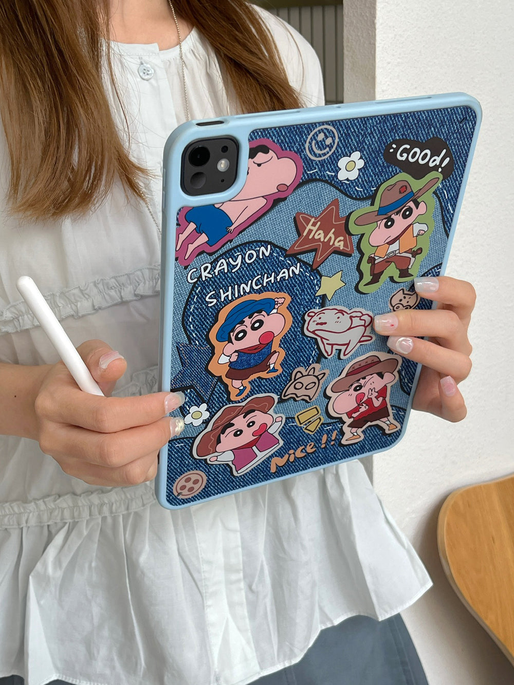 Side angle view of a pastel blue protective iPad case with Crayon Shin-chan cartoon graphics held by a person holding a white Apple Pencil against a soft indoor background.