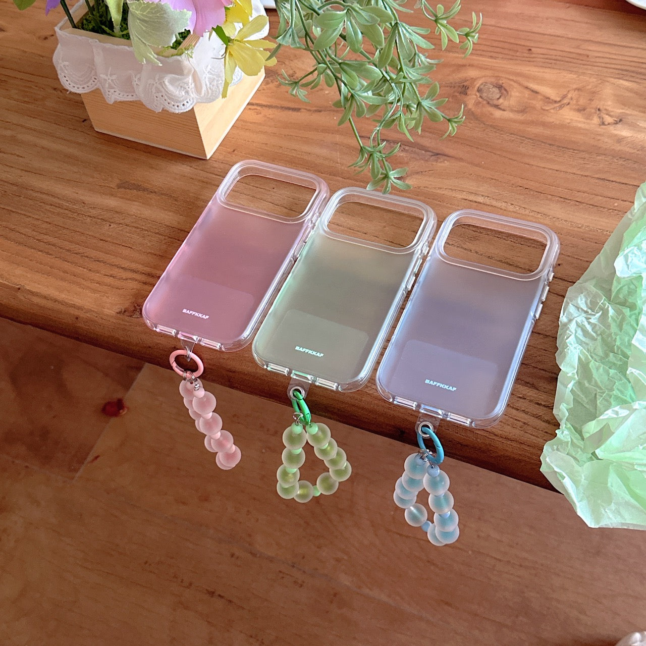 Three pastel-colored clear bumper phone cases in pink, green, and blue lined up on a wooden table with matching straps.