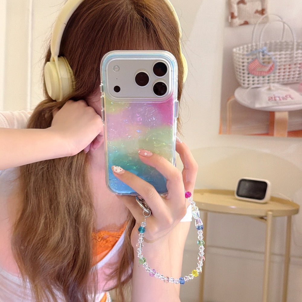 A girl with headphones taking a mirror selfie holding a pastel rainbow gradient iPhone case with an iridescent shell texture and a colorful crystal beaded wrist strap.