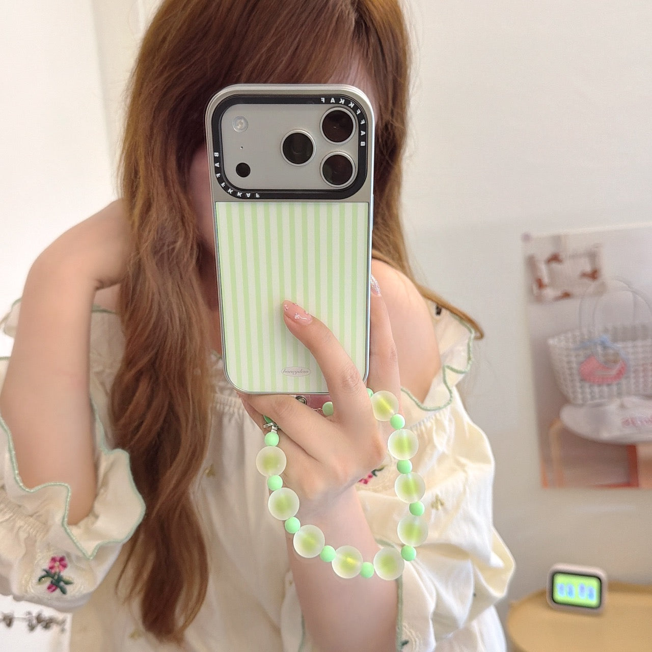 Mirror selfie showing a pastel green vertical stripe phone cover with a silver camera frame and beaded strap.
