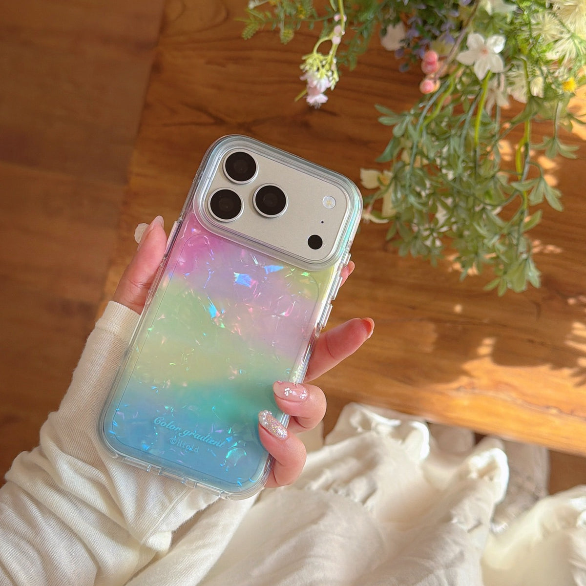 A hand showing a pastel pink to blue gradient shimmer iPhone case with an opal flake pattern, shot against a cozy cottagecore background.