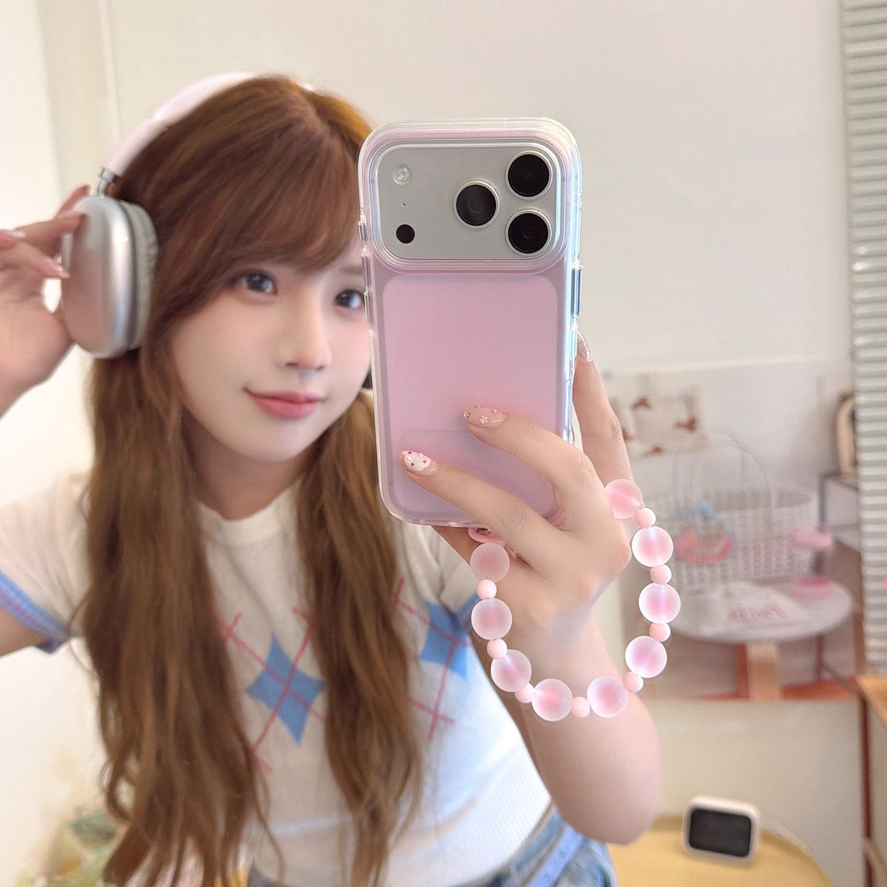 A young woman wearing headphones takes a mirror selfie with a pastel pink iPhone case and a matching frosted beaded wrist strap.