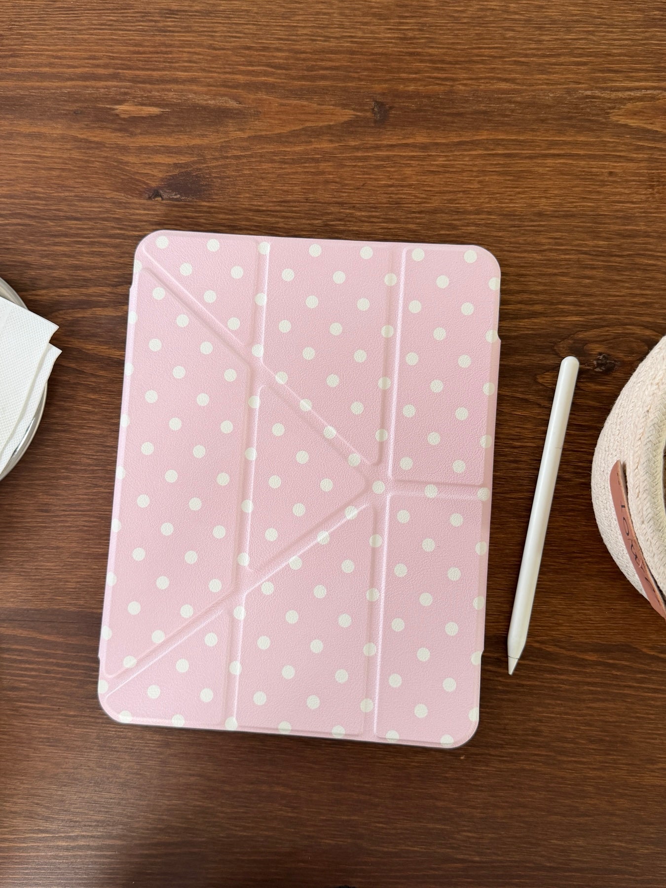 A top-down flat lay of a closed pastel pink polka dot ipad case placed on a dark wooden table next to a white Apple pencil.
