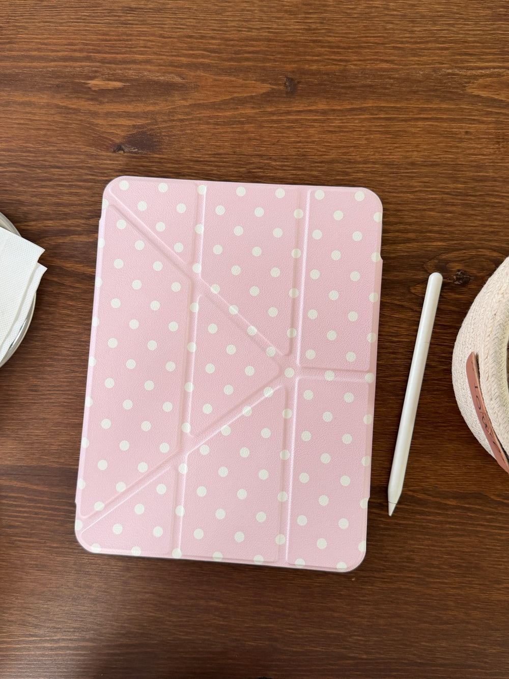 A top-down flat lay of a closed pastel pink polka dot ipad case placed on a dark wooden table next to a white Apple pencil.