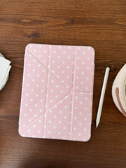 A top-down flat lay of a closed pastel pink polka dot ipad case placed on a dark wooden table next to a white Apple pencil.