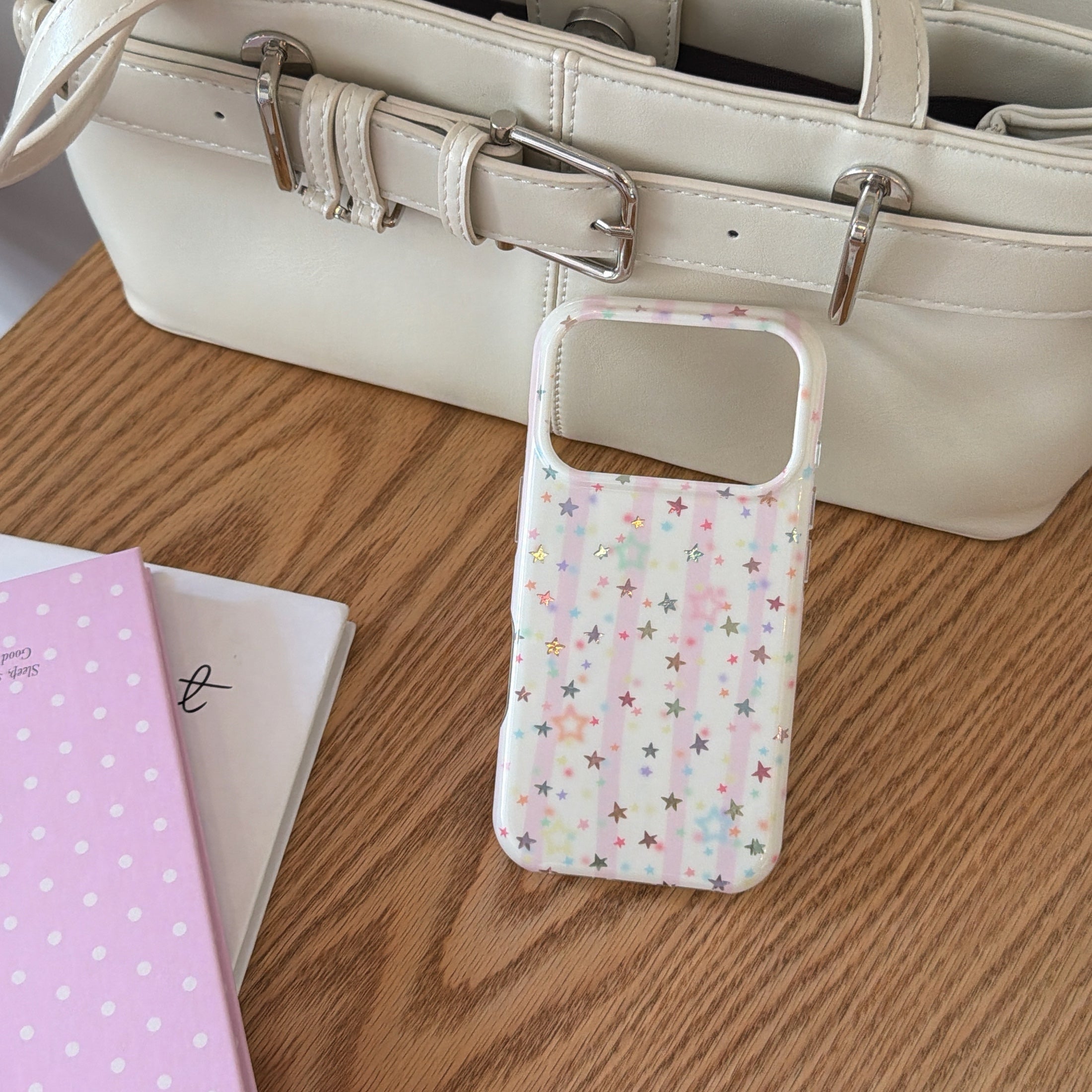 A pastel pink stripe phone case standing on a wooden desk next to a pink notebook and a white handbag.