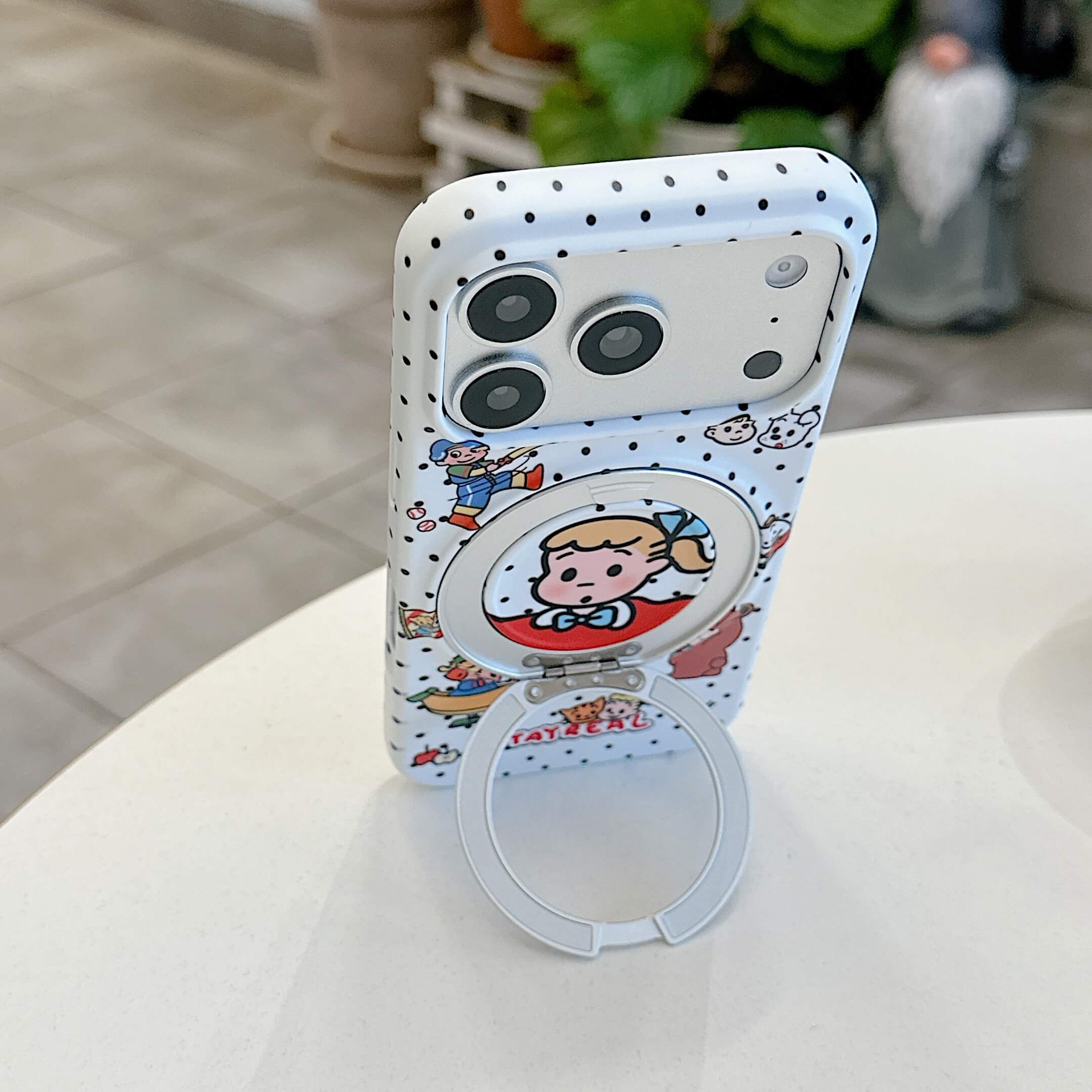 The cute polka dot cartoon iPhone case standing vertically on a table supported by its ring kickstand.