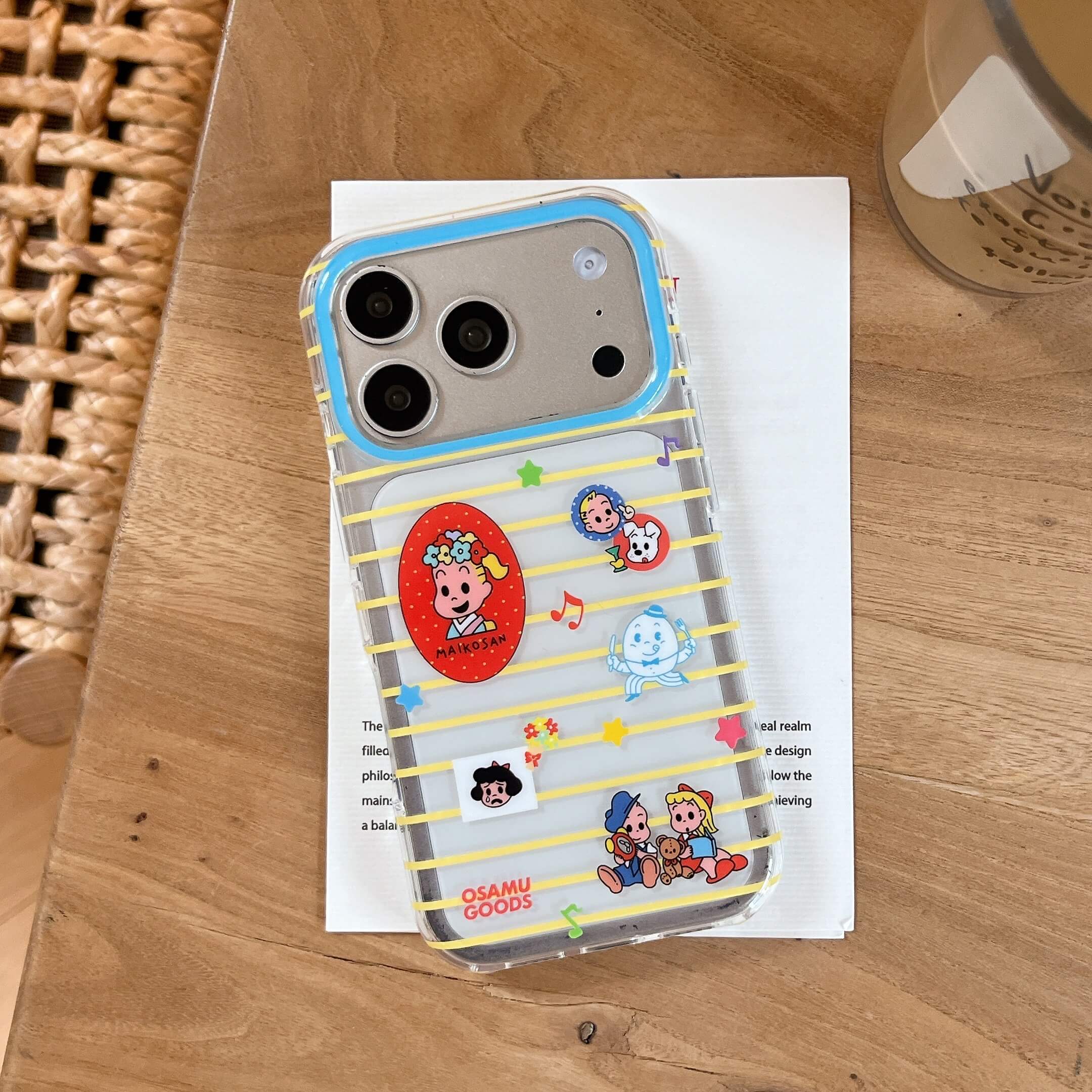 An Osamu Goods yellow striped iPhone case resting on a white paper over a wooden table next to a cup of coffee.